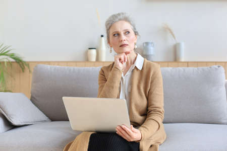 Cheerful middle aged woman using laptop while sitting on sofa at home