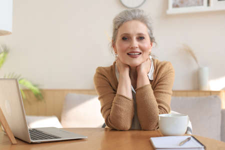 Middle aged woman sitting at a table with a laptop and looking at the camera smilingの写真素材