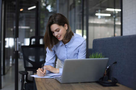 Smiling pretty woman sits at table. Happy entrepreneur writes notes to clients