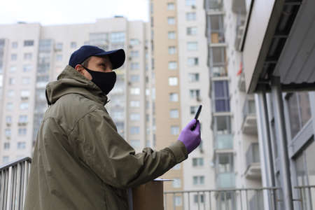 Courier in face mask with a parcel at the entrance of home verifies the information in smartphone
