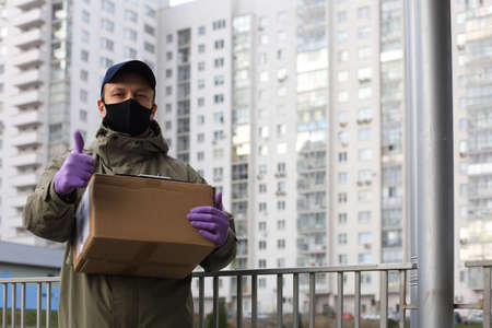Courier in medical mask and gloves with parcel stands near home outdoor, shows thumbs up....