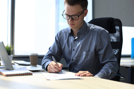 Portrait of handsome smiling man in casual shirt taking notes at workplaceの写真素材