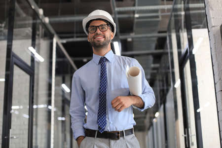Confident young business man in hardhat holding blueprint and looking away while standing indoors
