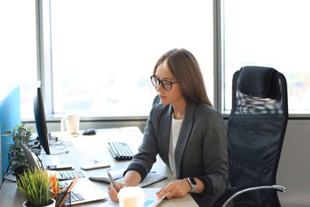 Beautiful business woman is writing something down while sitting in the office desk.の写真素材