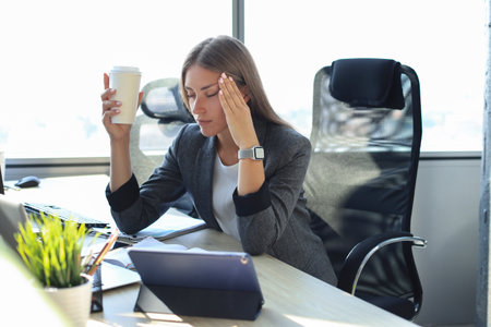 Image of business woman looking while tired while sitting at her working place and holding paper cup in handの写真素材