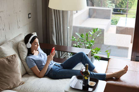 Pretty girl using her smartphone on couch at home in the living room. Listening music, drinking red wine, relaxation after a hard week at workの写真素材