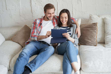 Young couple watching media content online in a tablet sitting on a sofa in the living roomの写真素材