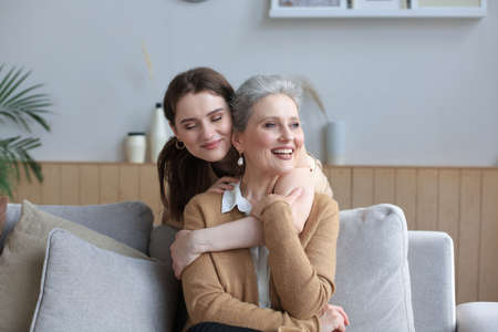 Happy daughter hugging older mother, standing behind sofa in living room, enjoying tender moment at...