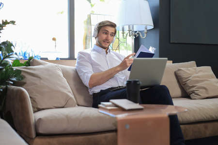 Concentrated young freelancer businessman sitting on sofa with laptop and examining documentsの写真素材
