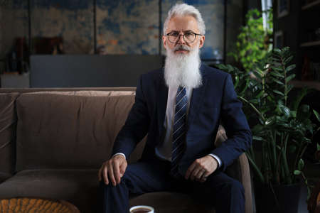Thoughtful confident handsome businessman thinking while sitting on sofa in his modern office about...