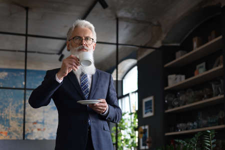 Start of a new working day with a cup of coffee. Handsome mature man holding coffee cup standing in his modern officeの写真素材