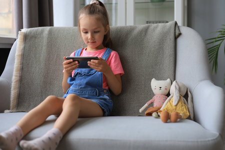 Smiling little girl sitting on sofa playing online games, web surfing information, using funny applications on smart phoneの写真素材
