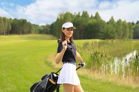 Woman in stylish summer golf outfit walking with bag of drivers on beautiful green course for golf gameの写真素材