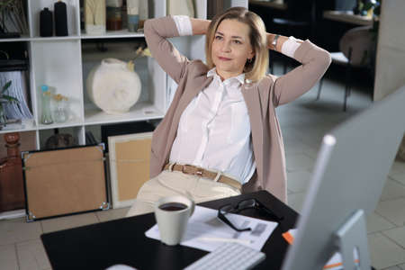 Woman with satisfied expression at desk. Arms up and folded behind her head in modern office.の写真素材