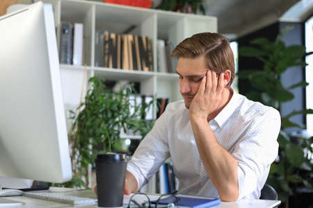 Tired young male sleeping sitting with computer behind desk at office
