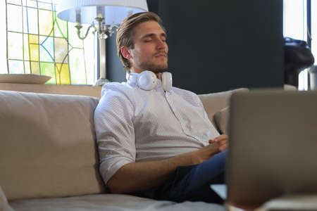 Tired young male sitting with closed eyes on sofa with laptop standing near him