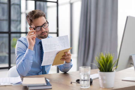 Handsome entrepreneur reading a letter from envelope in a desktop at office