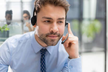 Cheerful young support phone male operator in headset, at workplace with collegues on the background while using computer, help service and client consulting call center conceptの写真素材