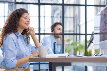 Female customer support operator with headset and smiling. Collegue is on the background