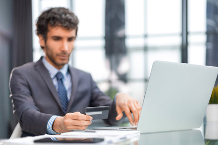 Smiling man sitting in office and pays by credit card with his laptopの写真素材