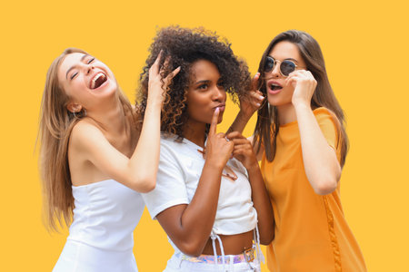 Three young smiling hipster women in summer clothes posing on yellow background. Female showing positive face emotionsの写真素材