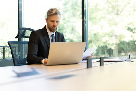 Mature businessman with beard working with documents, contracts and bills sitting at table using...