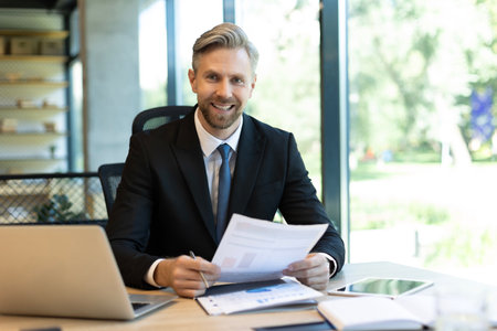 Mature businessman with beard working with documents, contracts and bills sitting at table using laptop at work in modern bright office with large windows.の写真素材