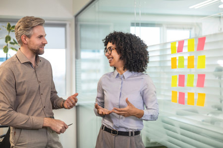 Two business people discussing work together using sticky notes on glass wall in a modern office