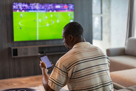 African man holding smartphone with sports bets app while watching football match at home in living...