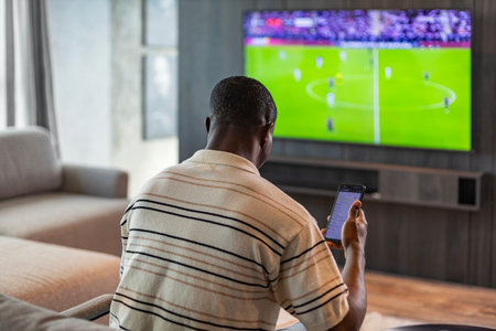 African man holding smartphone with sports bets app while watching football match at home in living room.の写真素材