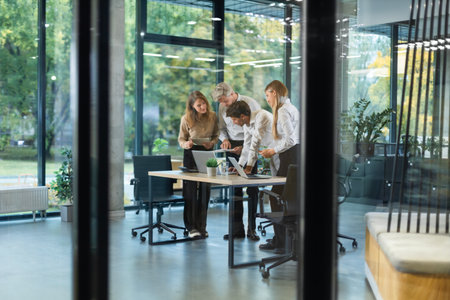 Group of business people standing together and discussing their work and projects, having a team meeting in an office, view from glass wall.の写真素材