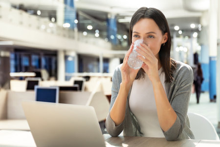 Positive caucasian businesswoman enjoying a refreshing break while drinking a glass of water, sitting at her desk in a modern office with a laptop.の写真素材