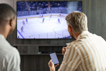 Two friends holding smartphone while watching ice hockey match at home in living roomの写真素材
