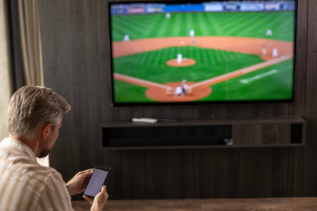 Caucasian man holding smartphone while watching baseball match at home in living room.
