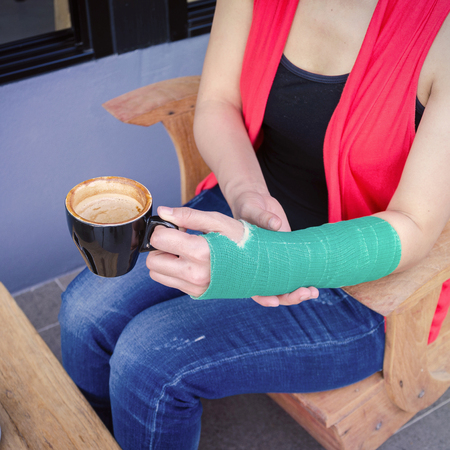 injured woman with green cast on the wrist holding black coffee cup and smart phones on wood table  in coffee cafe.の写真素材