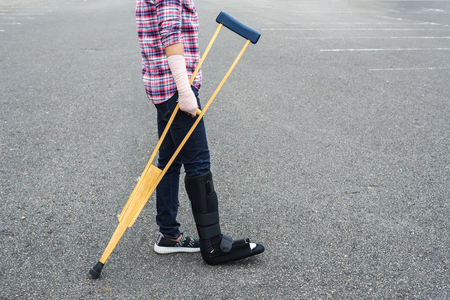 broken leg, injury woman wearing shirt and jeans  with arm splint and black leg splint standing on floor holding wooden crutches.の写真素材