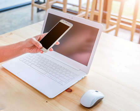 female hands using smart phones and working with a laptop in office at wooden desk, morning light included.の写真素材