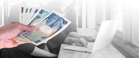 hand woman with Japanese currency yen bank notes on background two woman working on laptop in office.の写真素材