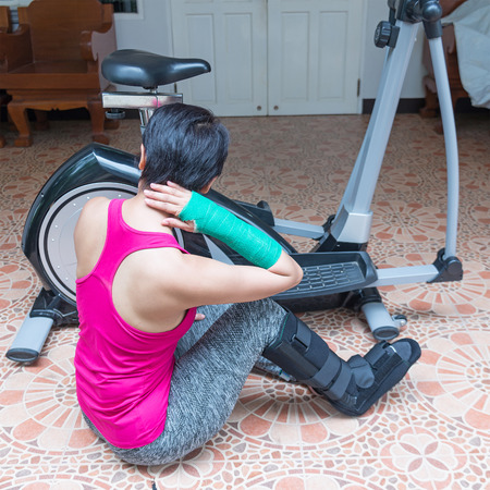 injured woman wearing sportswear with green cast on arm and black ankle brace on leg sitting on floor.の写真素材