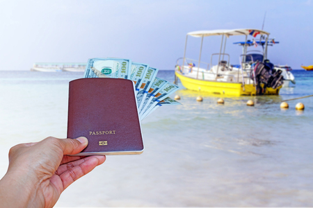 hand holding passport and American dollar currency isolated on blurred yellow boat and sea view, summer travel concept.の写真素材