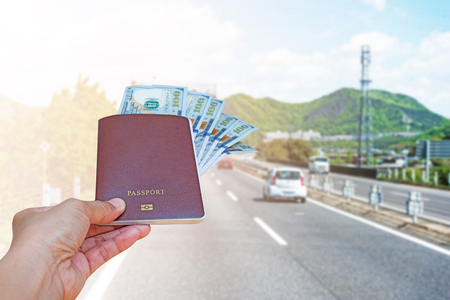 woman hand holding passport and American dollar currency isolated on blurred cars run on highway with mountain background.の写真素材