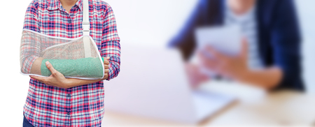 hand in a sling with broken arm in green cast isolated on blurred background working woman with office desk workplace with laptop and tablet in office.の写真素材