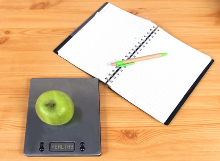 Open blank pages notebook with green apple on kitchen scale representing health diet lifestyle conceptの写真素材