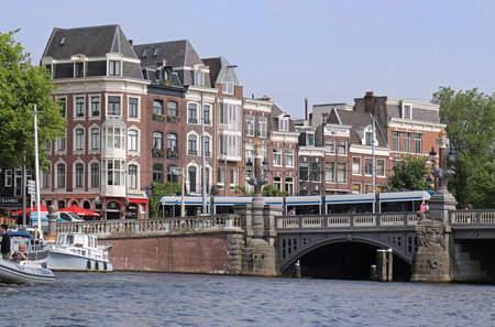 AMSTERDAM, NETHERLANDS - MAY 16, 2018: Cityscape of Blauwburg bridge with lantern poles in shape of crowns over Amstel river in Amsterdam with tram going across and people crossing the bridge by foot.のeditorial素材