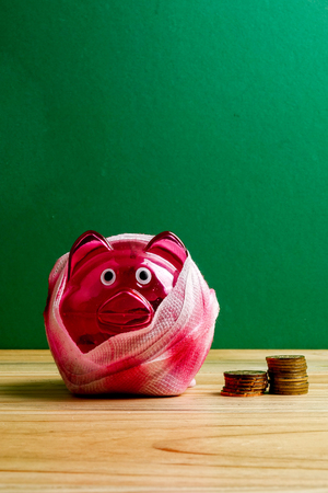 SAVING  DETERIORATE CONCEPT. Red piggy bank with bandage and small stack of coins on the wooden table over green background.の写真素材