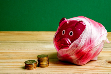 SAVING  DETERIORATE CONCEPT. Red piggy bank with bandage and small stack of coins on the wooden table over green background.の写真素材