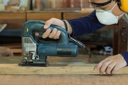 Carpenter is sawing a wooden plank with jig saw machine in carpentry workshop. He is wearing safety...
