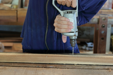 Hand of worker drills a hole with wooden plank using electric drill machine in workshopの写真素材