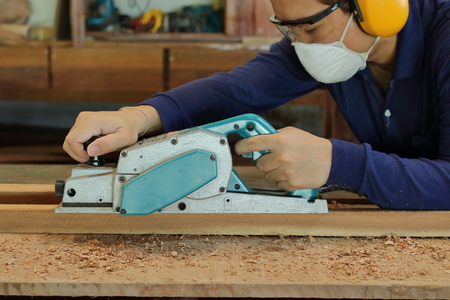 Carpenter using electric planer with wooden plank in carpentry workshop. He is wearing safety equipmentの写真素材