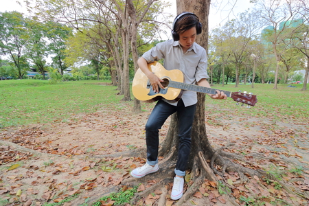 Wide angle shot. Guitarist man with headphones standing and playing acoustic guitar in outdoor park.の写真素材